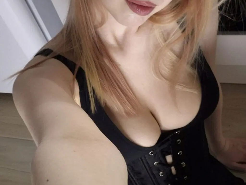 Becky Covert sits on a bed with long blonde hair, holding her breasts with both hands.