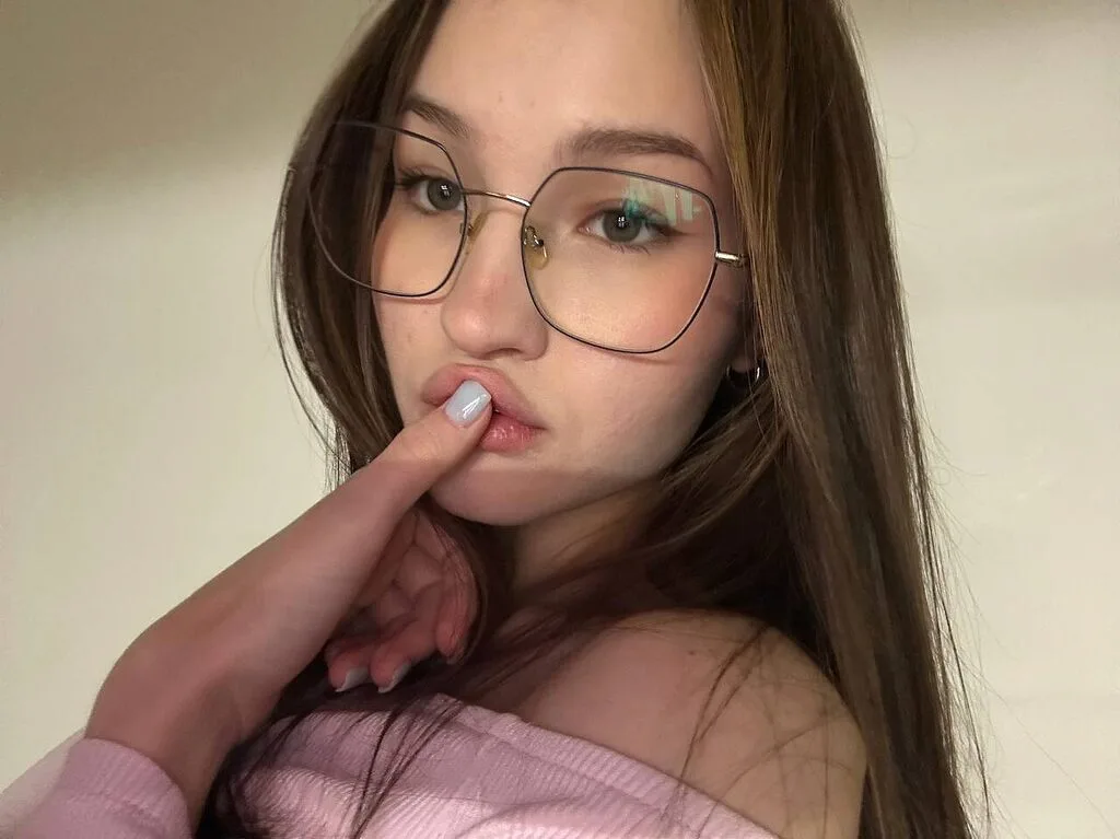 Kandi Zeff wears large glasses and a pink off-the-shoulder top while posing with her finger to her lips.