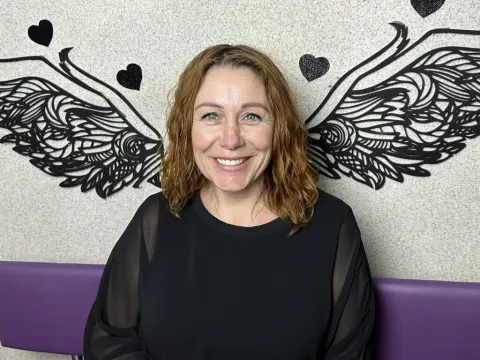 Taylor Foxtt — Taylor Foxtt smiles while sitting in front of a wall decorated with black angel wings and hearts.