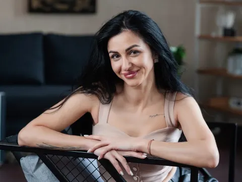 Eva Clarkk — Eva Clarkk smiles while leaning forward in a beige tank top with her long black hair flowing down.