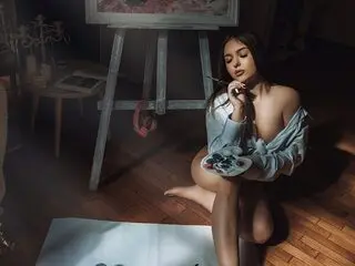 Lindsey Lu — Lindsey Lu sits on a wooden floor in a studio, wearing a loose light blue shirt and holding a paintbrush near her face.