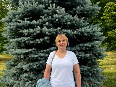 Kira Smiling — Kira Smiling stands in front of a large blue evergreen tree wearing a white v-neck t-shirt and denim shorts.