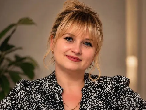 Kira Smiling — Kira Smiling smiles at the camera wearing a black and white patterned blouse with her blonde hair in a messy bun.
