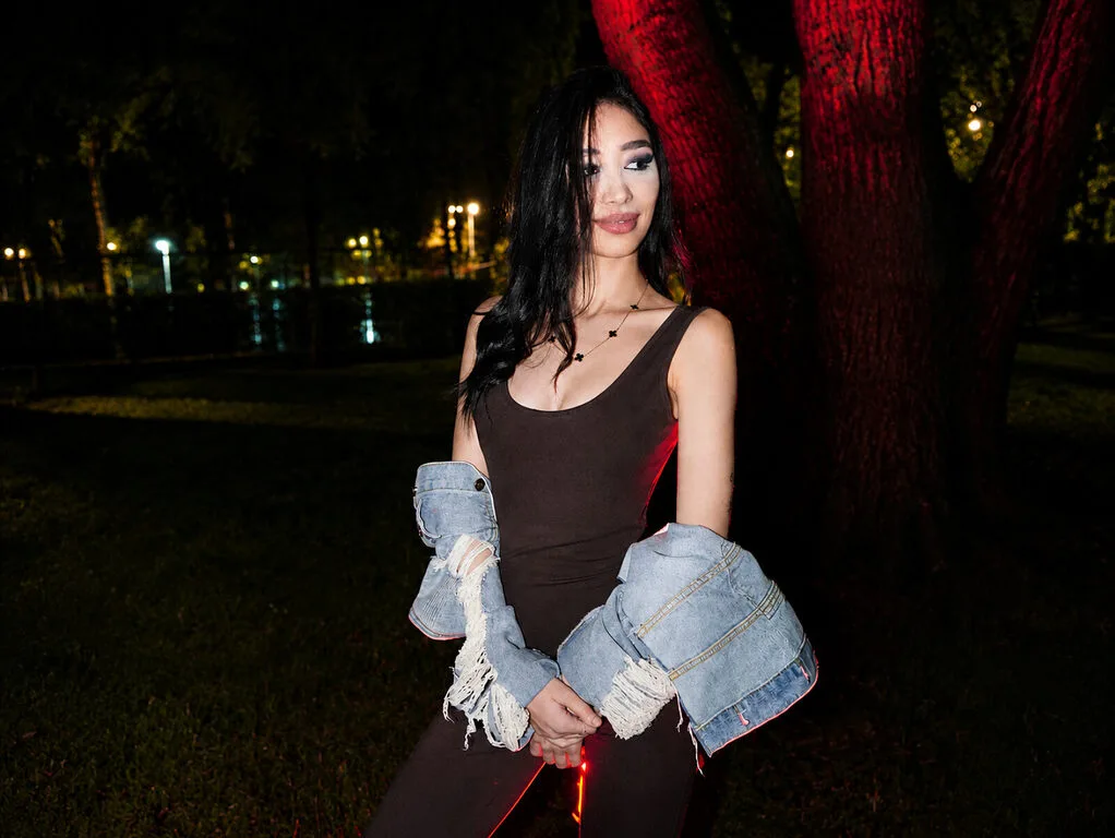 Lunaa Bliss wears a tight brown bodysuit and a denim jacket while posing outdoors at night.