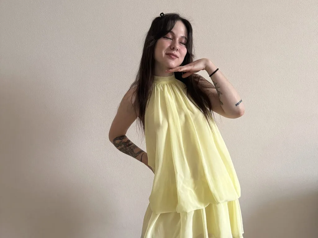 Holly Wallis poses with her eyes closed, wearing a light yellow sleeveless dress against a plain wall.