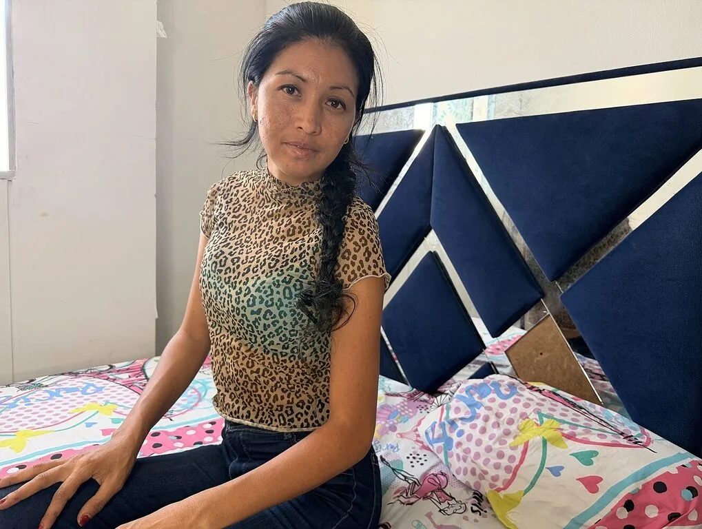 Emma Noir sits on a bed wearing a leopard print sleeveless top and dark jeans, leaning against a navy blue headboard.
