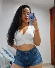 Luciana_Bloom poses in a white crop top and denim shorts, showing her long dark hair and cleavage while holding a phone for a mirror selfie.