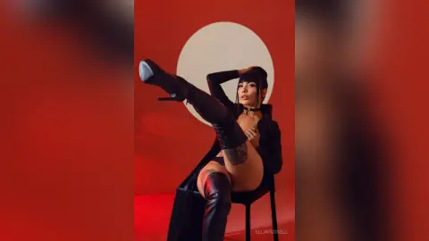 ElliaRussell poses in black lingerie and thigh-high boots, lifting one leg high against a red and white background.