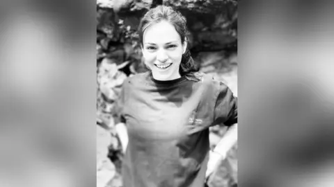 Smiling young woman in a casual t-shirt posing outdoors against a natural rocky background in black and white.