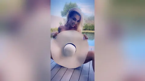 FelishaRose smiles while hiding her chest behind a large white sun hat on a wooden deck by a pool.