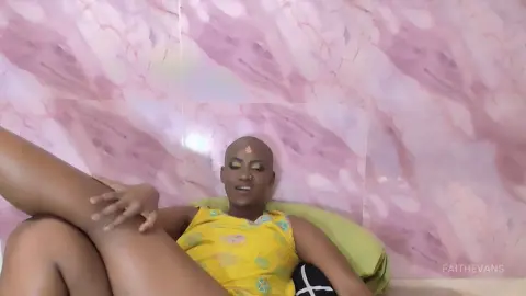 Smiling bald woman in a yellow patterned sleeveless top relaxing against a pink marble wall background.