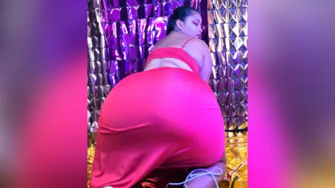 Curvy woman in a tight pink midi dress posing against a shimmering gold sequin backdrop with vibrant purple lighting.