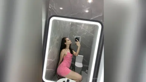 Young woman in a pink slip dress taking a selfie in a modern marble bathroom with a lighted mirror.