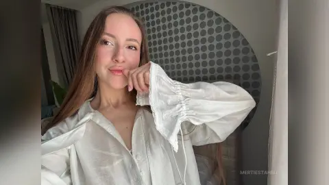Young woman in a white linen blouse posing in a bright, modern bedroom with a grey patterned wall.