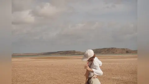 LaurentBelluci wears a white cap and a white long-sleeve shirt while standing in a vast, sandy desert landscape under a cloudy sky.