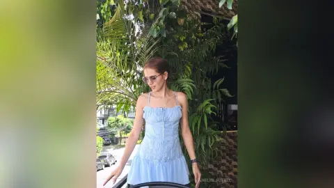 AmelieLautrec wearing a light blue sundress and sunglasses, standing outdoors next to green tropical plants and a brick wall.