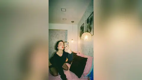 Young woman in a black outfit taking a moody selfie on a bed in a dimly lit bedroom.