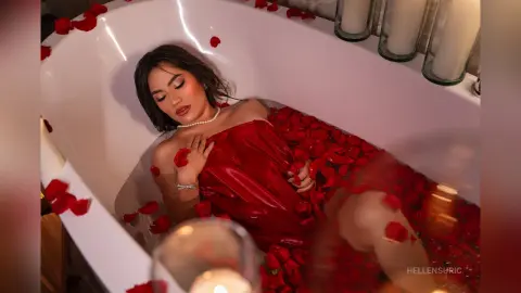 Glamorous woman in a red satin dress relaxing in a white bathtub filled with red rose petals.