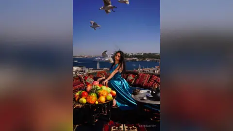 Woman in a blue satin dress sitting by a fruit platter on a rooftop overlooking a coastal city with flying seagulls.