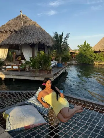 Woman in a yellow sundress relaxing on a wooden net hammock over tropical resort water at sunset.