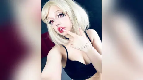 Blonde woman in black lingerie with dramatic eye makeup and red lips posing in a moody setting.