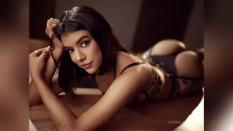 Beautiful woman in black lace lingerie posing on a wooden floor in a moody, dimly lit bedroom setting.