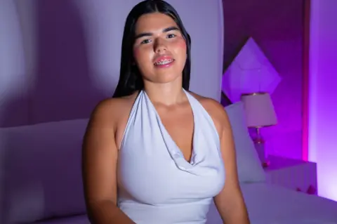 Smiling young woman in a white halter dress sitting on a bed in a moody, purple-lit bedroom.