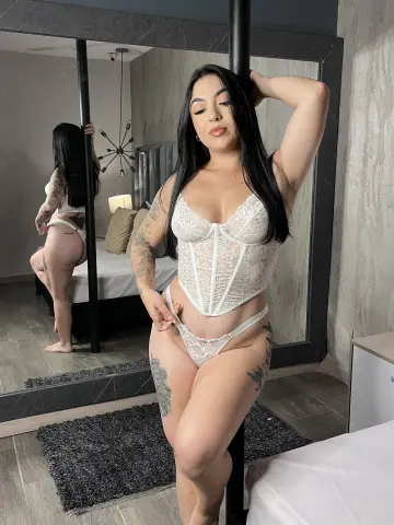 BellaFloyd poses in a white lace corset and matching thong, showing off her long black hair and tattoos while leaning against a black pole.