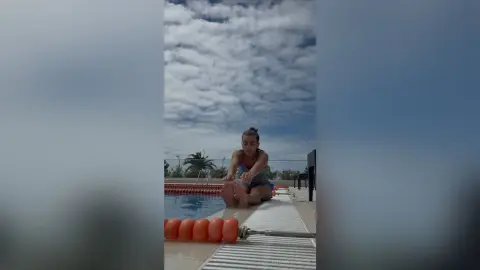 Young woman in a pink swimsuit sitting by a swimming pool under a cloudy sky.