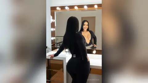 Smiling woman in a sleek black long-sleeve bodysuit posing in a brightly lit dressing room.