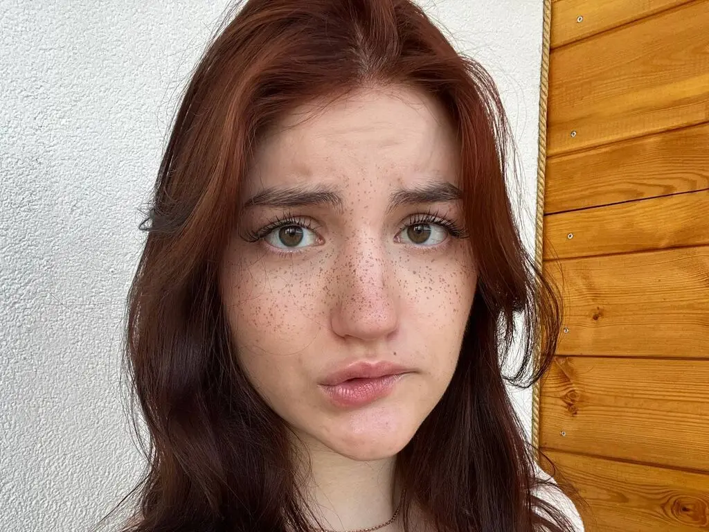 Close-up portrait of a freckled woman with long red hair looking thoughtfully against a white and wood wall.