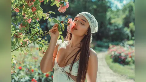 AlessiaCrush wears a white lace top and a headscarf while smelling a pink rose in a sunny garden.