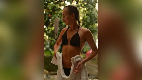 Smiling woman in a black bikini top holding a white towel in a lush green outdoor garden setting.