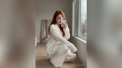 Redheaded woman in white striped pajamas sitting by a bright window in a minimalist modern room.