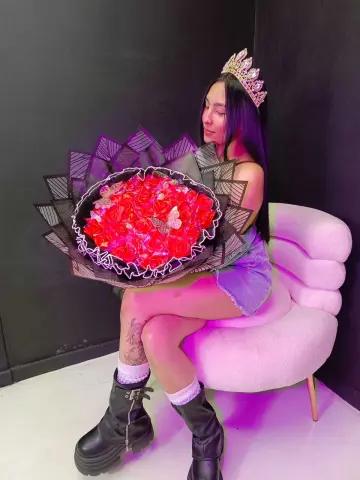 MorganHarper wearing a silver crown and denim shorts, sitting in a white chair while holding a large bouquet of red roses.