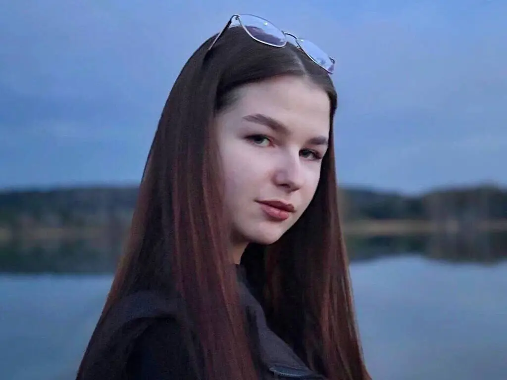 Young woman with long brown hair and sunglasses looking calm by a serene lake at dusk.