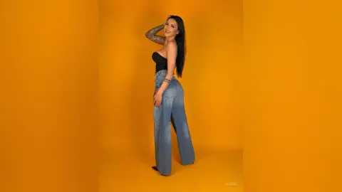 Smiling woman in a black tube top and blue wide-leg jeans posing against a vibrant yellow studio background.
