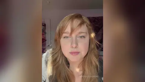 Redheaded woman with nose ring in casual grey clothing looking calmly at the camera in a bright indoor setting.