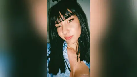 Young woman with black bangs in a blue denim shirt posing for a moody indoor selfie.