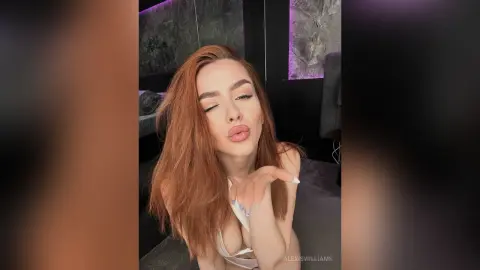 AlexisWilliams with long red hair pouts her lips while wearing a white bra in a dark room.