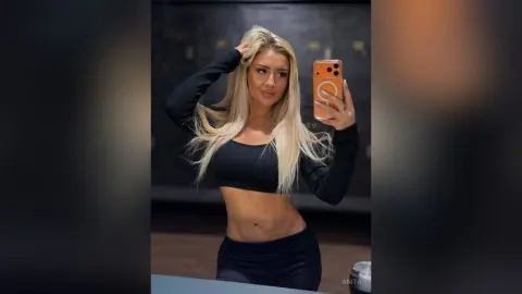Anita poses for a mirror selfie in a black long-sleeve crop top and black leggings, running her hands through her long blonde hair.