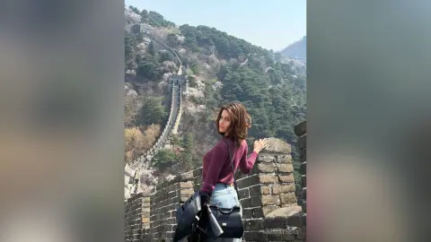 Young woman in a purple sweater standing by the Great Wall of China on a sunny day.