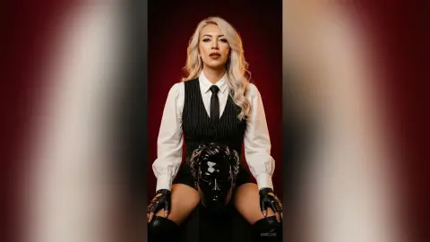 Blonde woman in a white shirt and black pinstripe vest posing confidently against a dark red background.