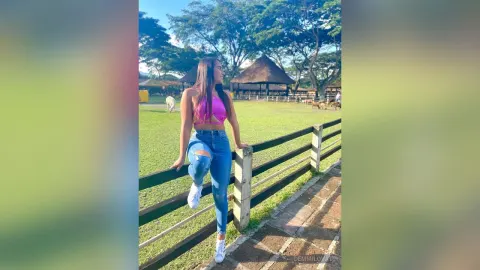 DemmiLovat — DemmiLovat leans against a wooden fence in a pink crop top and ripped blue jeans outdoors.