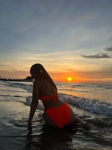 Woman in orange bikini sitting in ocean waves during a golden sunset at the beach.