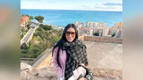 Aleja smiles wearing sunglasses and a black patterned scarf while overlooking a coastal city and the ocean.