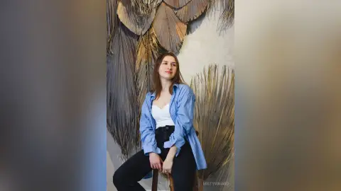 Young woman in a blue striped shirt and black pants sitting against a textured neutral wall background.
