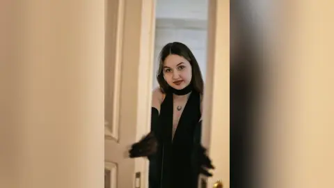 Young woman in a chic black outfit and choker posing elegantly in a bright doorway.