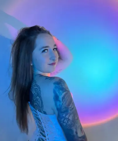 Tattooed woman in a white corset posing under dreamy blue and pink neon lighting.