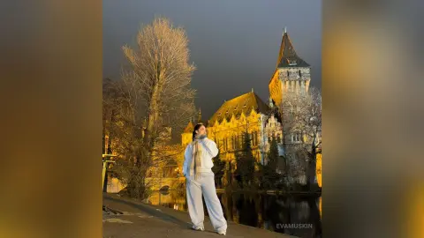 EvaMuskin stands outdoors at night wearing white pants and a white top, posing in front of a large lit building and trees.